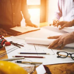 Zespół osób analizuje plan architektoniczny na drewnianym stole, widoczne narzędzia pomiarowe, ołówek, okulary i kask BHP w rogu kadru.