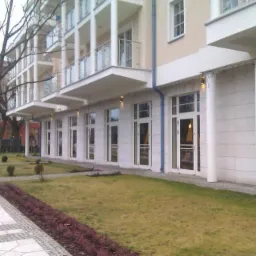 Elewacja budynku z białymi oknami i drzwiami, widok z zewnątrz na trawnik i chodnik.