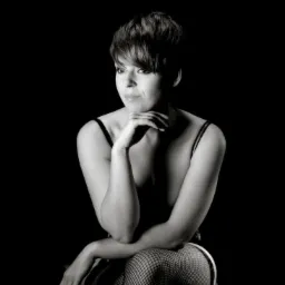 Czarno-biały portret kobiety z krótkimi włosami, oparta broda na dłoni, siatka na nogach, ciemne tło.