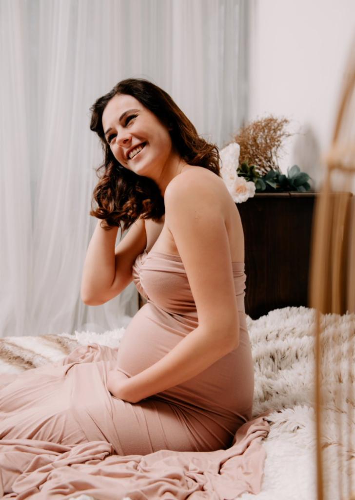 Uśmiechnięta kobieta w beżowej sukni ciążowej siedzi na futrzanym łóżku, trzymając dłonie na brzuchu, z dekoracją kwiatową w tle.