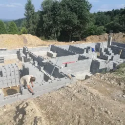 Budowa fundamentów domu z bloczków betonowych, pracownik w jasnej koszulce układa bloczki, widoczne zbrojenia i wykop z ziemią w tle.