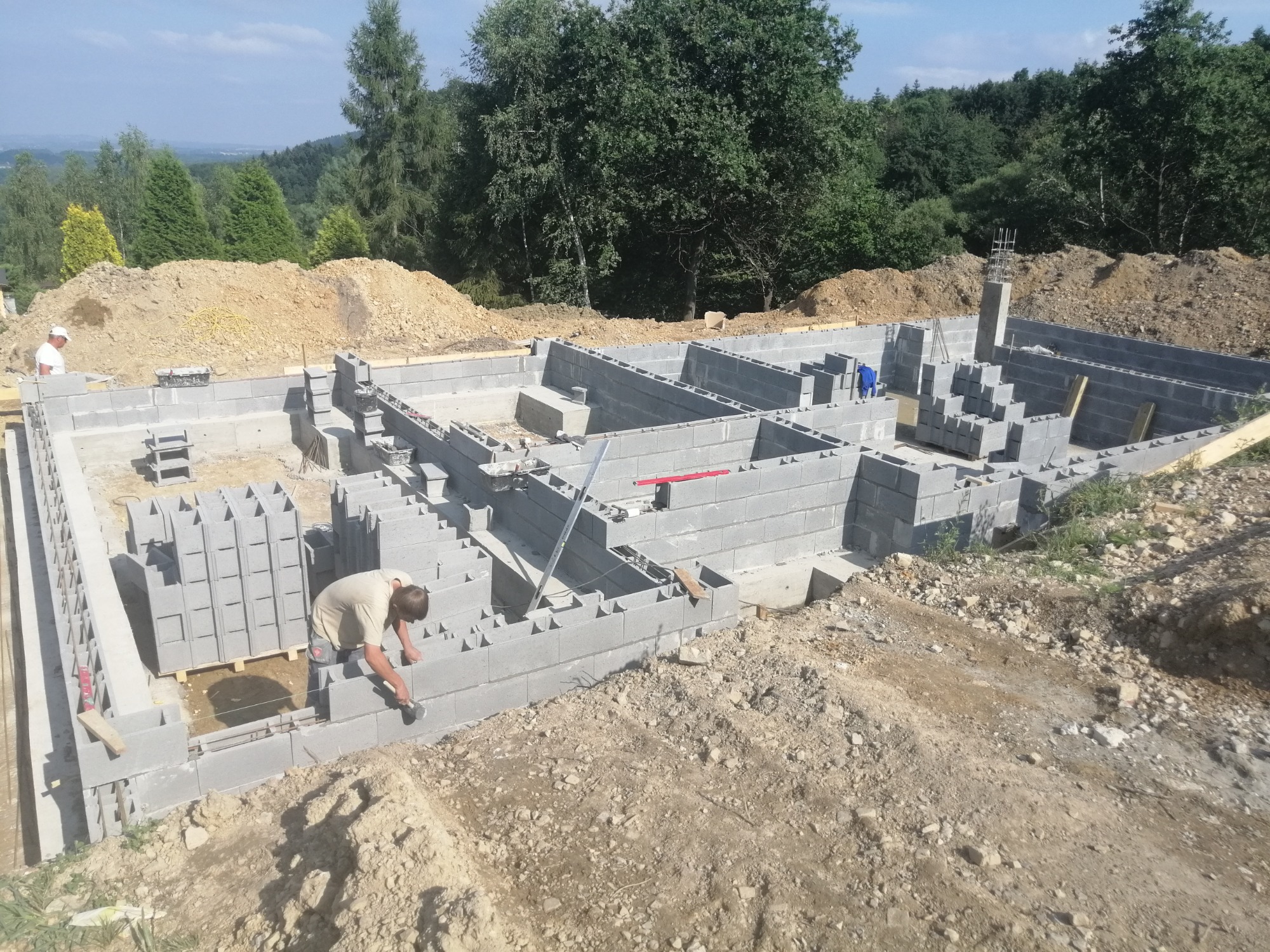 Budowa fundamentów domu z bloczków betonowych, pracownik w jasnej koszulce układa bloczki, widoczne zbrojenia i wykop z ziemią w tle.