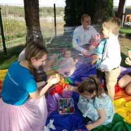 Dzieci malują farbami na folii przymocowanej do drzewa, pod nadzorem dorosłych, na tle kolorowej tkaniny w plenerze.