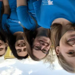 Cztery uśmiechnięte kobiety w niebieskich koszulkach z logo firmy Pracownia rozwoju i kreatywności, stojące blisko siebie na zewnątrz, z budynkiem i drzewami w tle.