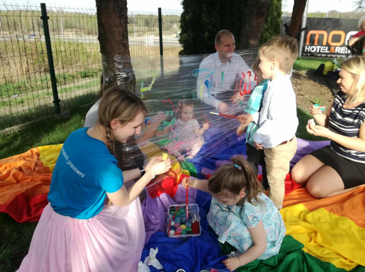 Dzieci malują farbami na folii przymocowanej do drzewa, pod nadzorem dorosłych, na tle kolorowej tkaniny w plenerze.