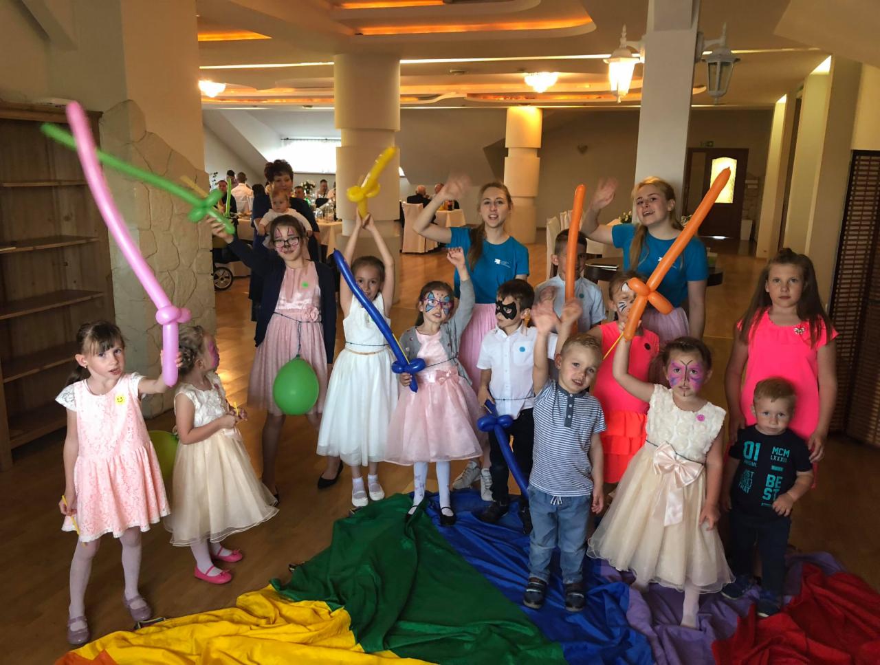 Grupa dzieci z wymalowanymi twarzami i balonami w kształcie mieczy, pozuje do zdjęcia na tle kolorowej tkaniny, podczas imprezy w sali z kolumnami.