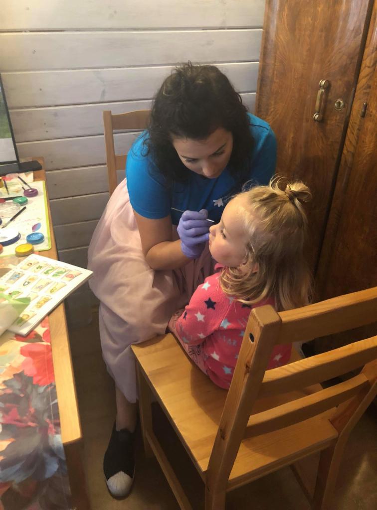 Kobieta w niebieskiej koszulce i fioletowych rękawiczkach maluje twarz małej dziewczynce siedzącej na drewnianym krześle, używając cienkiego pędzelka i farb umieszczonych na stole obok.