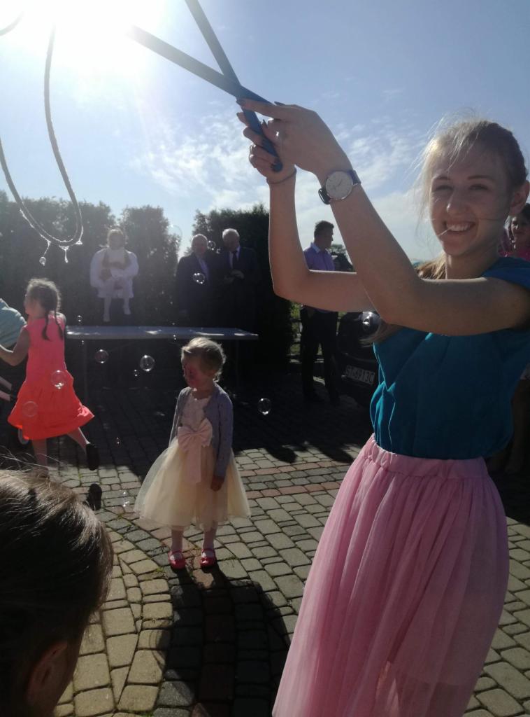 Uśmiechnięta kobieta w turkusowej bluzce i różowej tiulowej spódnicy puszcza bańki mydlane dużym przyrządem, w tle dzieci w sukienkach i grupa mężczyzn w garniturach, prawdopodobnie podczas...