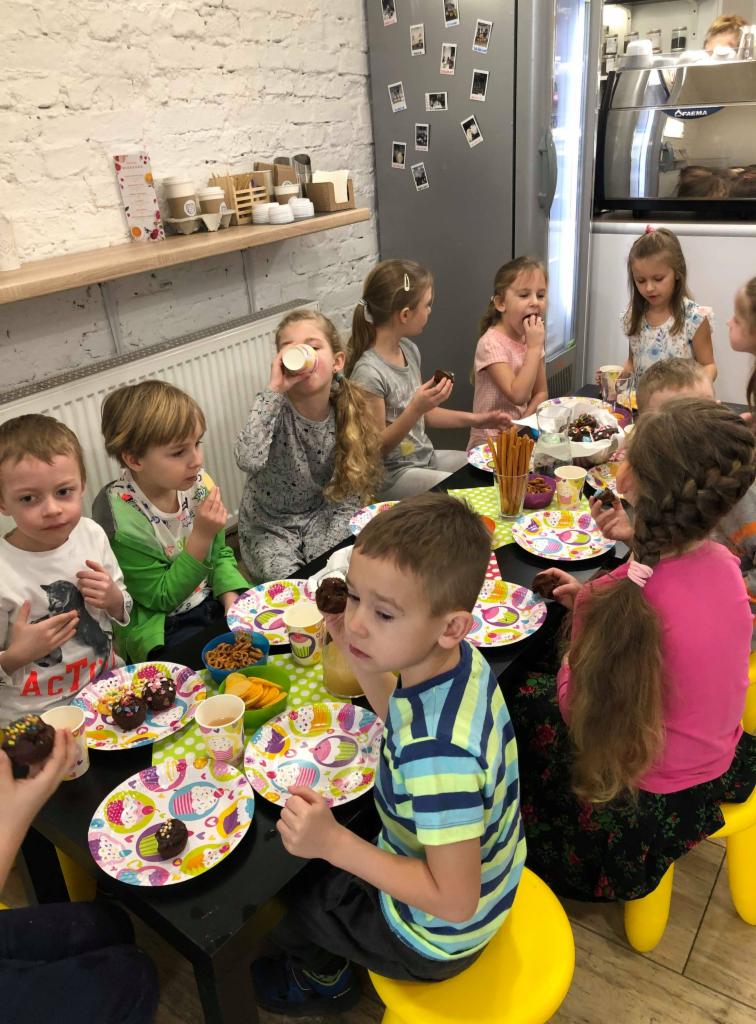 Grupa dzieci siedzi przy stole, jedząc babeczki i pijąc napoje podczas przyjęcia; widoczne kolorowe talerzyki i dekoracje.
