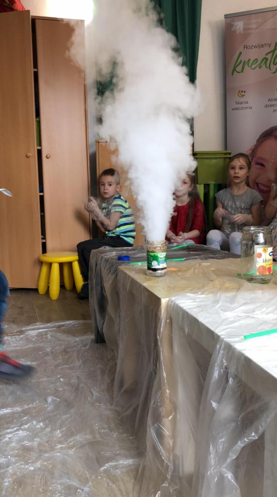 Dzieci obserwują eksperyment z gęstym białym dymem wydobywającym się ze słoika na stole zabezpieczonym folią, w tle szafa i plakat z napisem 'Rozwijamy kreatywność'.