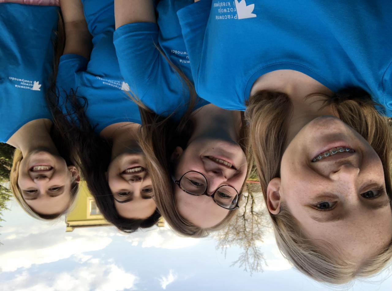 Cztery uśmiechnięte kobiety w niebieskich koszulkach z logo firmy Pracownia rozwoju i kreatywności, stojące blisko siebie na zewnątrz, z budynkiem i drzewami w tle.