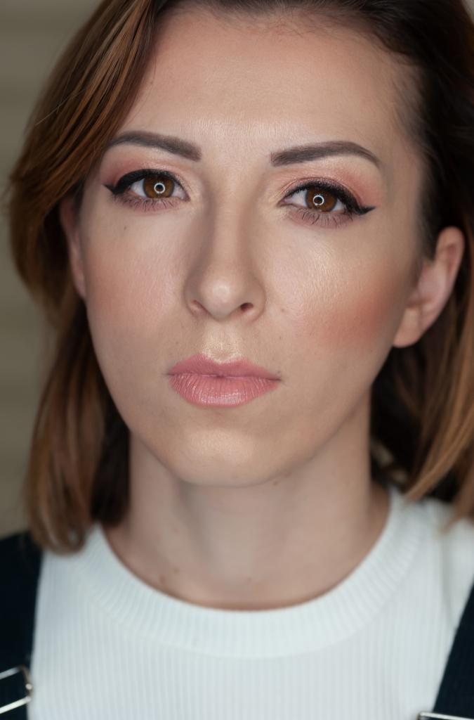 Portret kobiety z delikatnym, dziennym makijażem: różowe cienie, czarna kreska na powiece, podkreślone kości policzkowe, błyszczące usta w odcieniu nude.