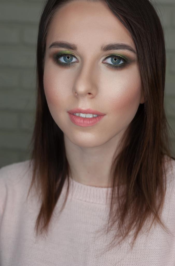 Portret kobiety z zielonym cieniowaniem powiek, różową szminką i delikatnym różem na policzkach, na tle ceglanej ściany.