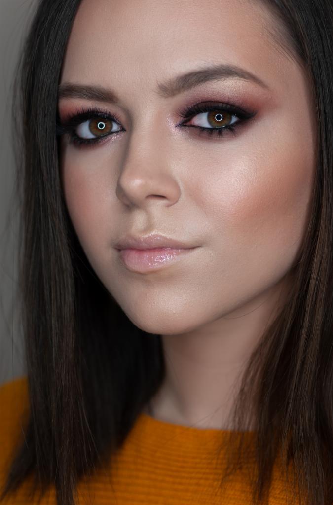 Portret kobiety z intensywnym, wieczorowym makijażem oczu w odcieniach różu i brązu, podkreślonymi brwiami i błyszczącymi ustami, w pomarańczowym swetrze.