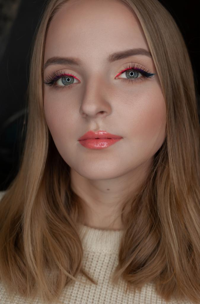 Portret kobiety z delikatnym, rozświetlonym makijażem, z neonowo-różową kreską na powiece i błyszczącymi, koralowymi ustami, włosy blond opadające na ramiona.