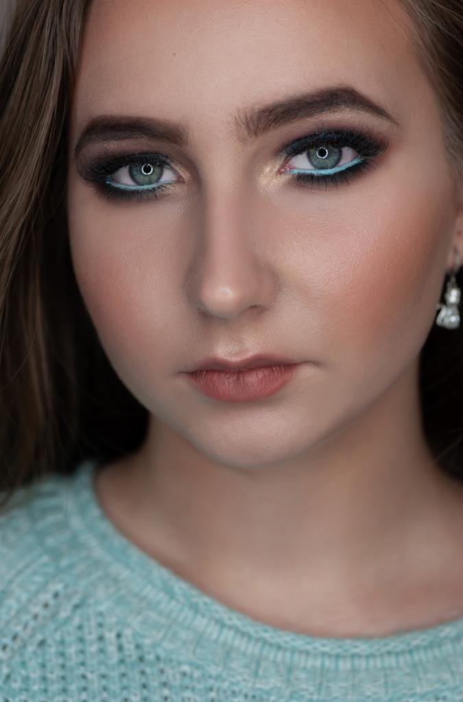 Portret kobiety z wyrazistym makijażem oczu: ciemne cienie, podkreślone dolne powieki turkusową kreską, brwi starannie wystylizowane, delikatny róż na policzkach i subtelna szminka.