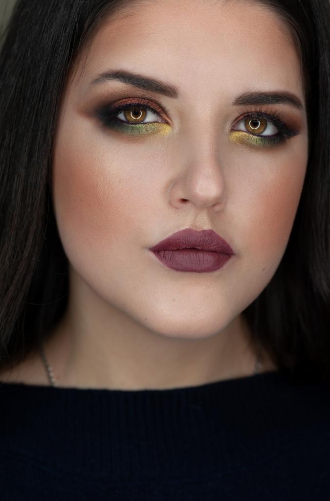 Portret kobiety z intensywnym, artystycznym makijażem oczu w odcieniach zieleni, brązu i złota oraz matową szminką w kolorze burgundu. Widoczne precyzyjne konturowanie twarzy i delikatny rozświetlacz.