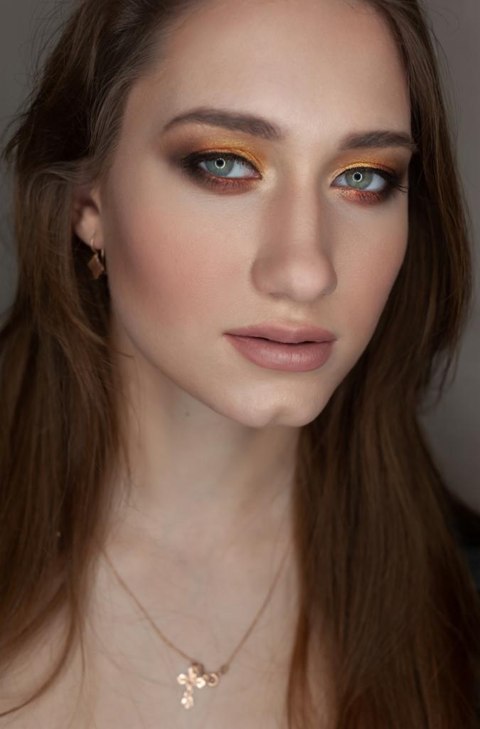 Portret kobiety z profesjonalnym makijażem oczu w odcieniach złota i brązu, delikatny makijaż ust, subtelna biżuteria, naturalne oświetlenie.