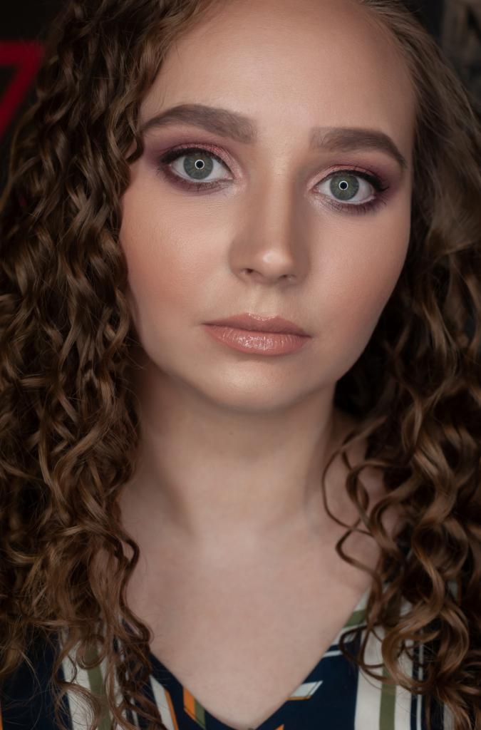 Portret kobiety z delikatnym makijażem oczu w odcieniach różu i brązu, podkreślającym zielone tęczówki; naturalny makijaż ust i burza brązowych loków.