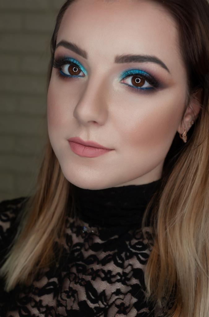 Portret kobiety z intensywnym, turkusowym cieniem do powiek, mocno wytuszowanymi rzęsami i delikatnym różem na policzkach, w czarnej koronkowej bluzce.