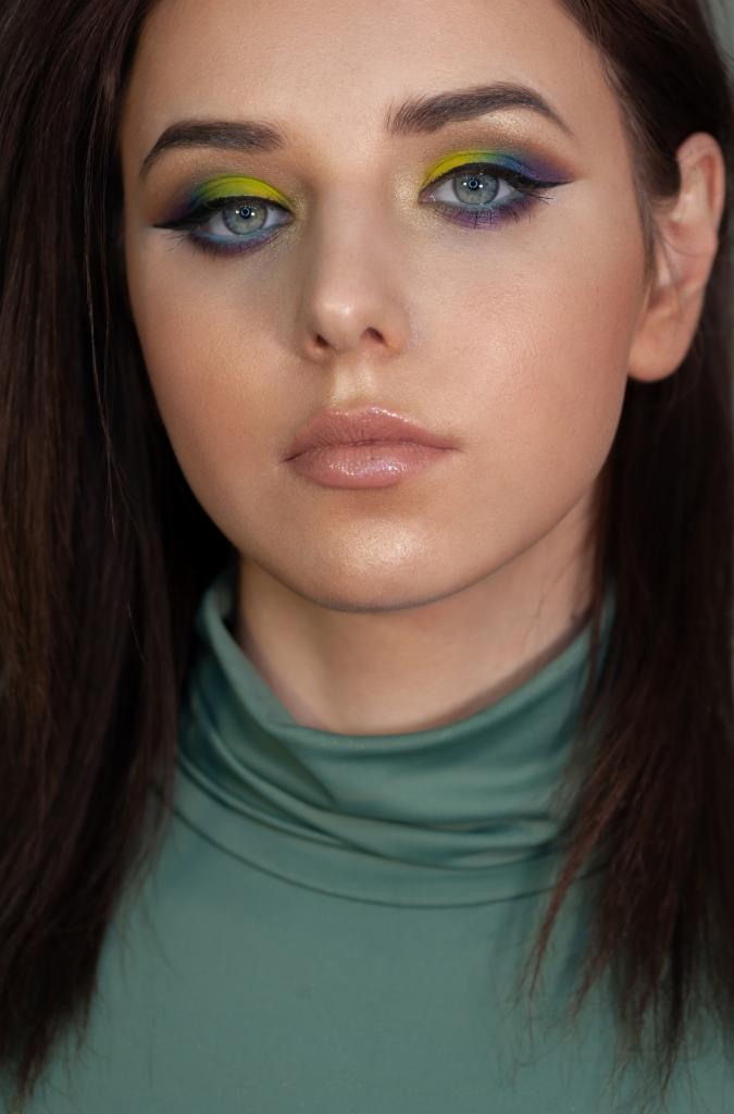 Portret kobiety z intensywnym, kolorowym makijażem oczu w odcieniach żółci, zieleni i fioletu, podkreślonym czarnym eyelinerem i błyszczącymi ustami w neutralnym odcieniu, ubrana w zielony golf.