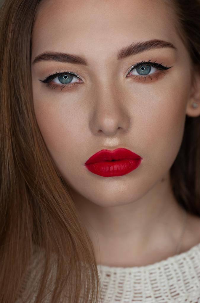 Portret młodej kobiety z intensywnym czerwonym kolorem ust, precyzyjną czarną kreską na powiekach i subtelnym srebrnym akcentem w wewnętrznym kąciku oka, podkreślającym jasnoniebieski kolor tęczówki.