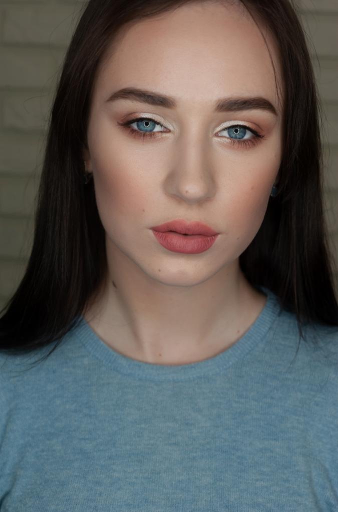 Portret kobiety z delikatnym, dziennym makijażem, podkreślone oczy z jasnym cieniem i subtelny róż na ustach, na tle jasnej ściany z cegieł.