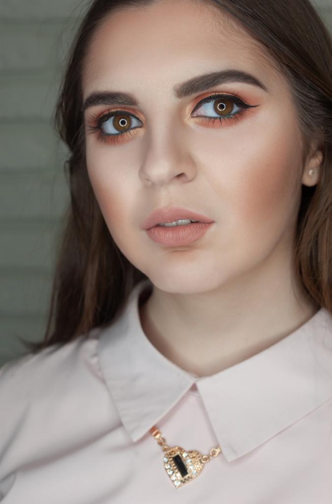 Portret kobiety z delikatnym, brzoskwiniowym cieniem na powiekach, brązowym eyelinerem i matową pomadką w odcieniu nude, uzupełniony subtelnym różem na policzkach i błyszczącym naszyjnikiem...