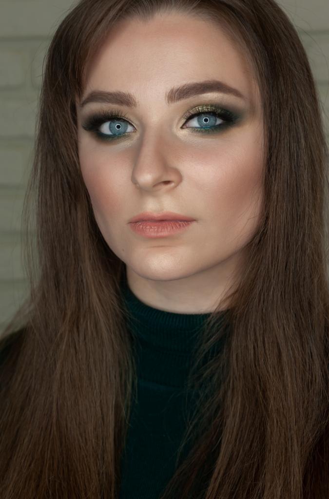 Portret kobiety z zielono-złotym makijażem oczu i długimi, prostymi, brązowymi włosami w ciemnozielonym golfie.