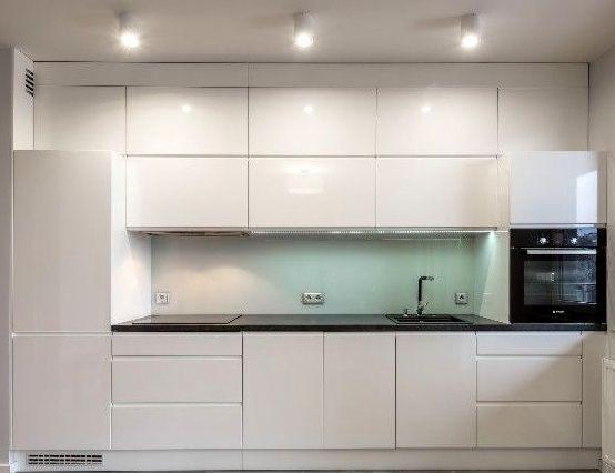 Nowoczesna, biała kuchnia na wymiar w Częstochowie z lakierowanymi frontami, czarnym blatem i szklanym panelem między szafkami, oświetlona punktowymi lampami sufitowymi.