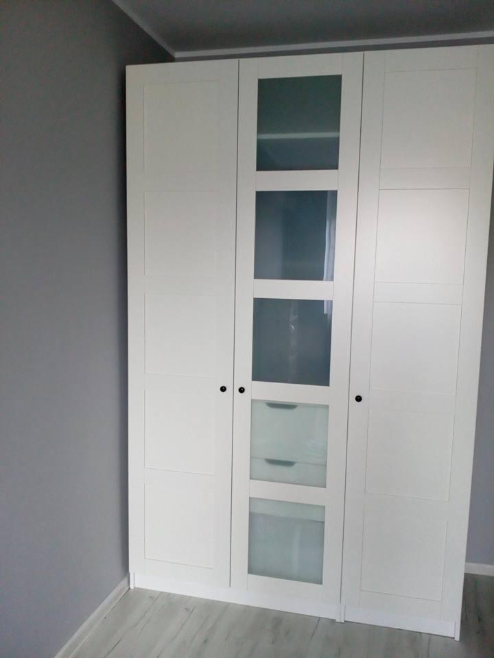 Trzydrzwiowa biała szafa z centralnymi drzwiami z szybami, widoczne półki i szuflady wewnątrz, na tle szarej ściany i podłogi imitującej drewno.