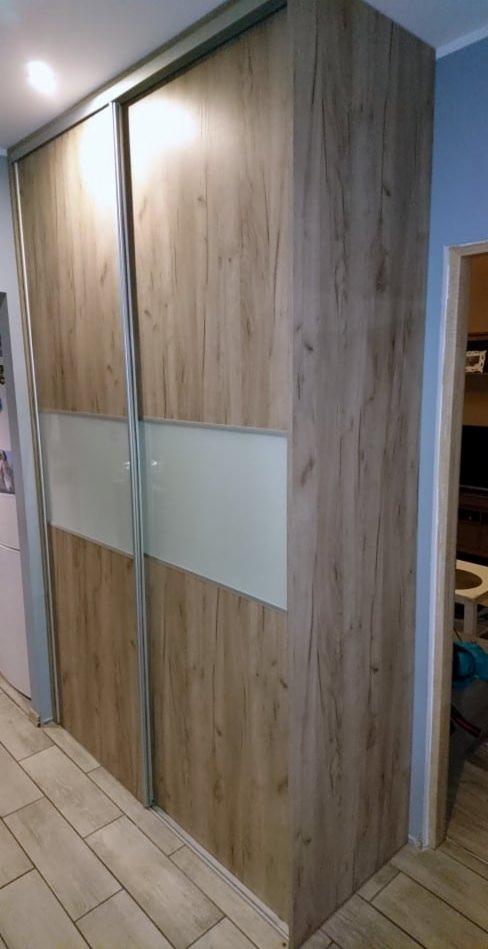 Dwudrzwiowa szafa wnękowa z przesuwnymi drzwiami, wykończona okleiną imitującą jasne drewno z poziomym pasem z matowego szkła, widoczna z boku na tle podłogi z płytek imitujących drewno.