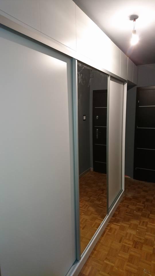 Biała szafa przesuwna z lustrem w zabudowie, widoczna podłoga parkietowa i drzwi w tle.
