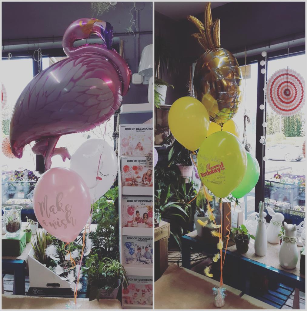 Dwa bukiety balonów: jeden z flamingiem, drugi z ananasem, otoczone roślinami w doniczkach i dekoracjami okiennymi.