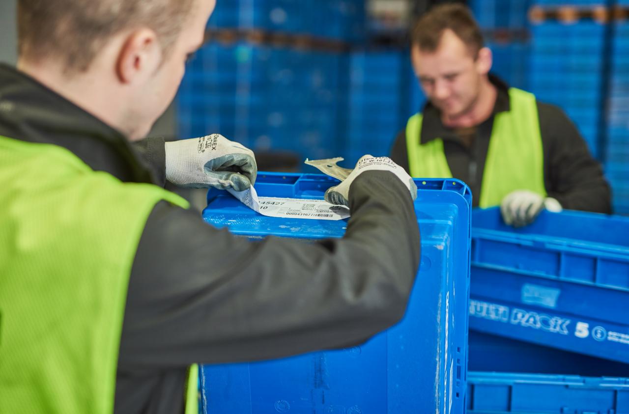 Pracownik w Rybniku etykietuje niebieski pojemnik w hali przemysłowej, drugi pracownik w tle, obaj w kamizelkach odblaskowych.