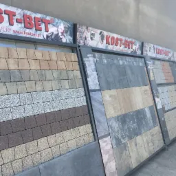 Ekspozycja różnych wzorów i kolorów kostki brukowej na stojakach z logo producenta Kost-Bet, prezentowanych na zewnątrz budynku.