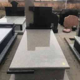 Nowoczesny, szary pomnik z czarnym krzyżem, odbijający światło w polerowanej powierzchni, ustawiony na placu wystawowym z innymi gotowymi elementami.