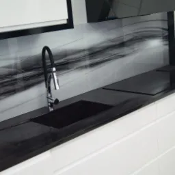 Czarny, połyskujący blat z wpuszczonym zlewem i płytą grzewczą na tle białych szafek kuchennych i panelu dekoracyjnego z motywem abstrakcyjnych szarości.