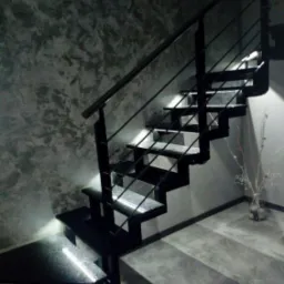 Nowoczesne schody kamienne z czarną metalową balustradą i podświetleniem LED, na tle ściany z efektem betonu architektonicznego.