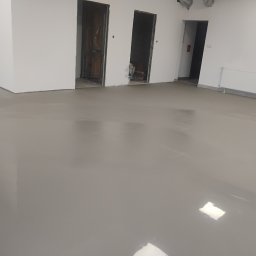 PQF DOSKONAŁE PODŁOGI - Wyrównana, gładka, szara wylewka samopoziomująca w nowym biurze. Widoczne przygotowane otwory drzwiowe i grzejnik. Podłoże gotowe do dalszych prac wykończeniowych.