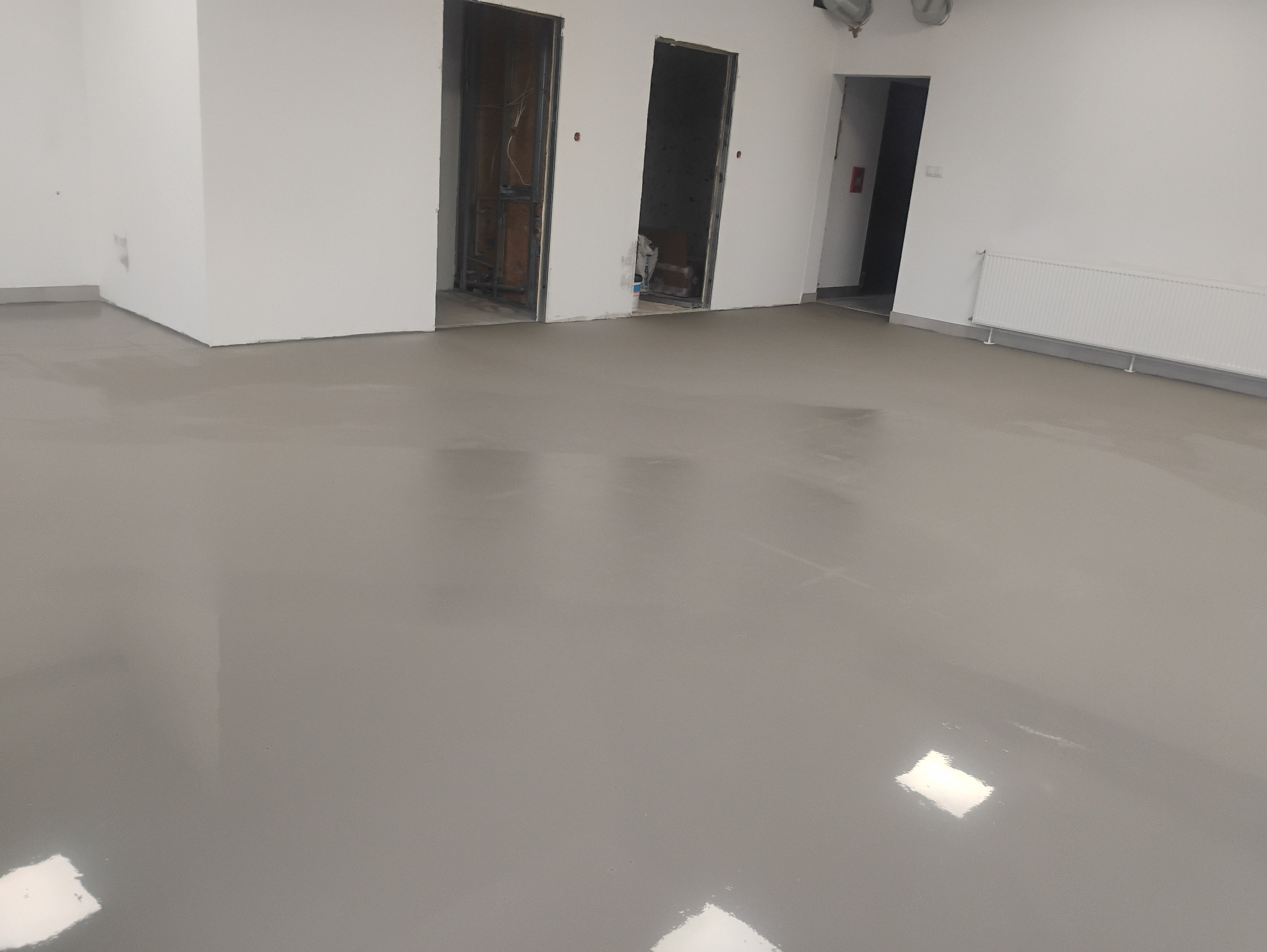 Wyrównana, gładka, szara wylewka samopoziomująca w nowym biurze. Widoczne przygotowane otwory drzwiowe i grzejnik. Podłoże gotowe do dalszych prac wykończeniowych.
