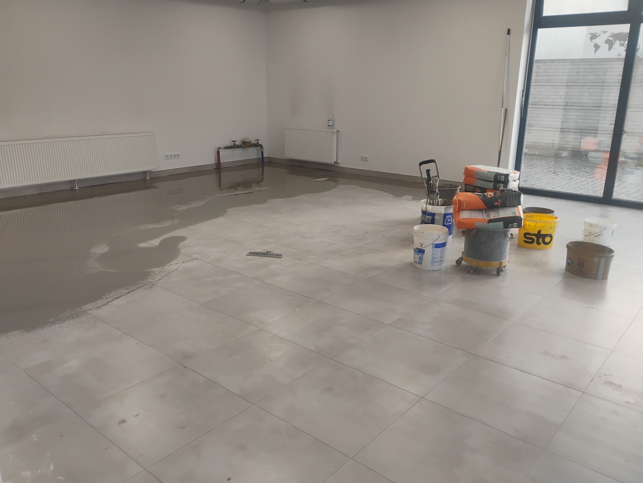 Wylewka samopoziomująca w trakcie prac w pomieszczeniu z grzejnikami i dużym oknem. Widoczne narzędzia i materiały budowlane, część podłogi już zalana. Szary odcień.