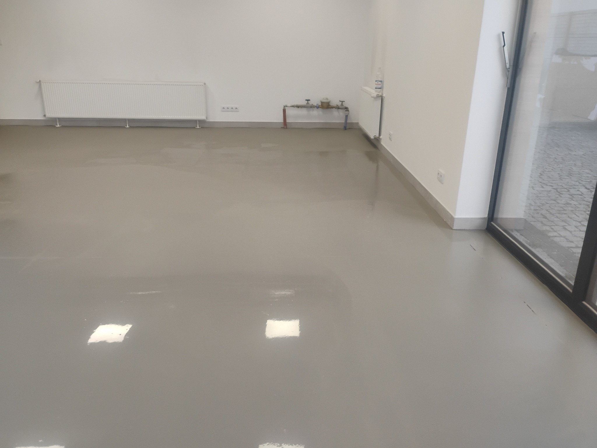 Jasnoszara, gładka posadzka w pustym pomieszczeniu biurowym, z grzejnikiem i instalacją wodną przy ścianie. Widoczne odbicia światła na podłodze i fragment okna.