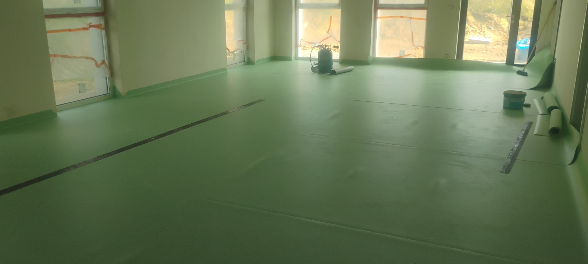 Wnętrze z przygotowaną, zieloną podłogą pod montaż wykładziny, widoczny sprzęt i materiały. Okna zabezpieczone folią, ściany w jasnym odcieniu. Przygotowanie do prac wykończeniowych.