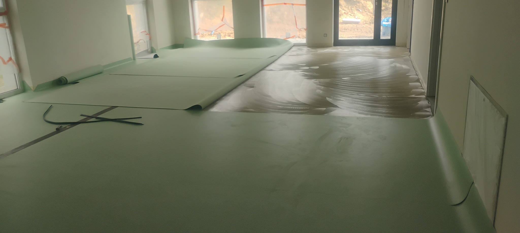 Wnętrze w trakcie prac: zielona wykładzina rozłożona częściowo na podłodze z wylanym podkładem. Rolka wykładziny i narzędzia leżą na powierzchni. Widoczne okna i drzwi.