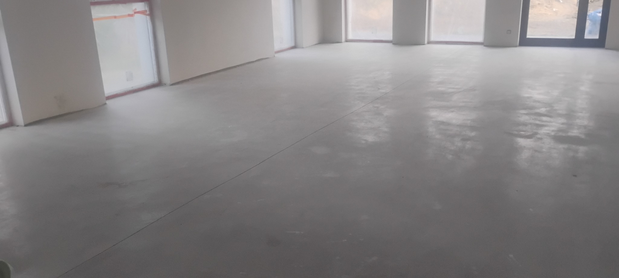 Wnętrze w stanie deweloperskim z wylaną, szarą posadzką. Widoczne okna i drzwi balkonowe. Delikatne, naturalne światło dzienne.