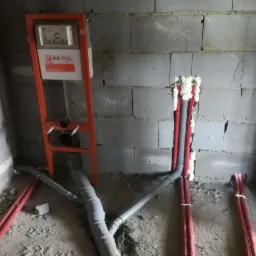 Surowy stan łazienki w trakcie montażu stelaża podtynkowego WC, widoczne rury kanalizacyjne z szarego PCV oraz czerwone rury doprowadzające wodę, zabezpieczone pianką montażową w bruzdach ściany...