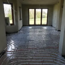 Białe rury ogrzewania podłogowego ułożone na folii z czerwoną siatką w surowym wnętrzu domu jednorodzinnego, widok przez przeszklone drzwi balkonowe na zielony ogród.