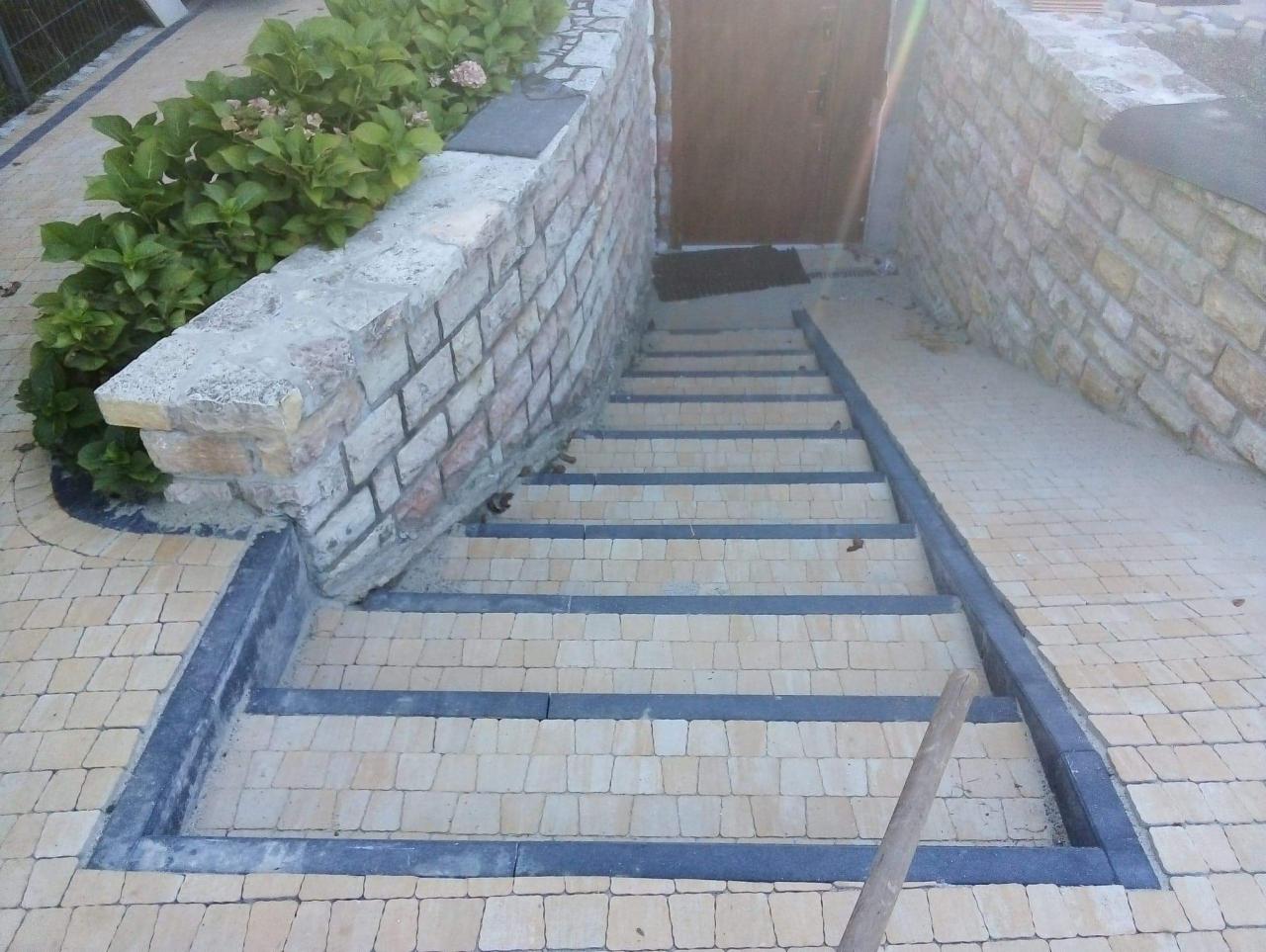 Schody zewnętrzne wyłożone jasną kostką brukową z ciemnymi obrzeżami, prowadzące do drzwi wejściowych, z kamiennym murkiem po lewej stronie obsadzonym hortensjami.