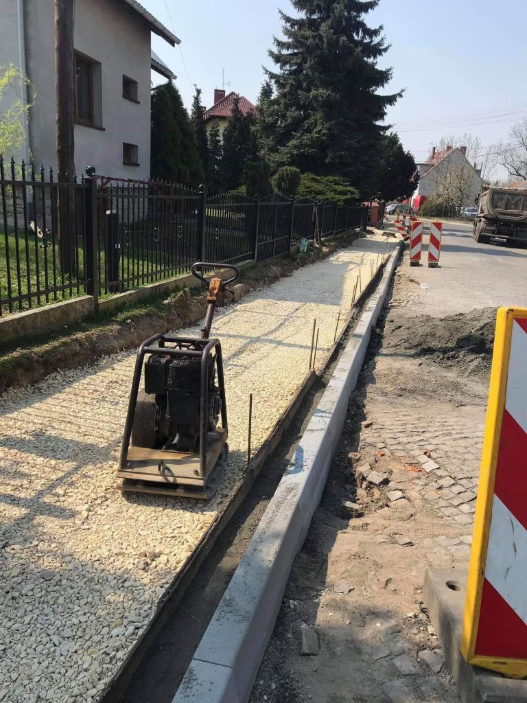 Przygotowanie podłoża pod chodnik: ubijarka wibrująca zagęszcza warstwę jasnego kruszywa wzdłuż krawężnika. Widoczne wystające pręty zbrojeniowe i fragment ulicy z kostką.